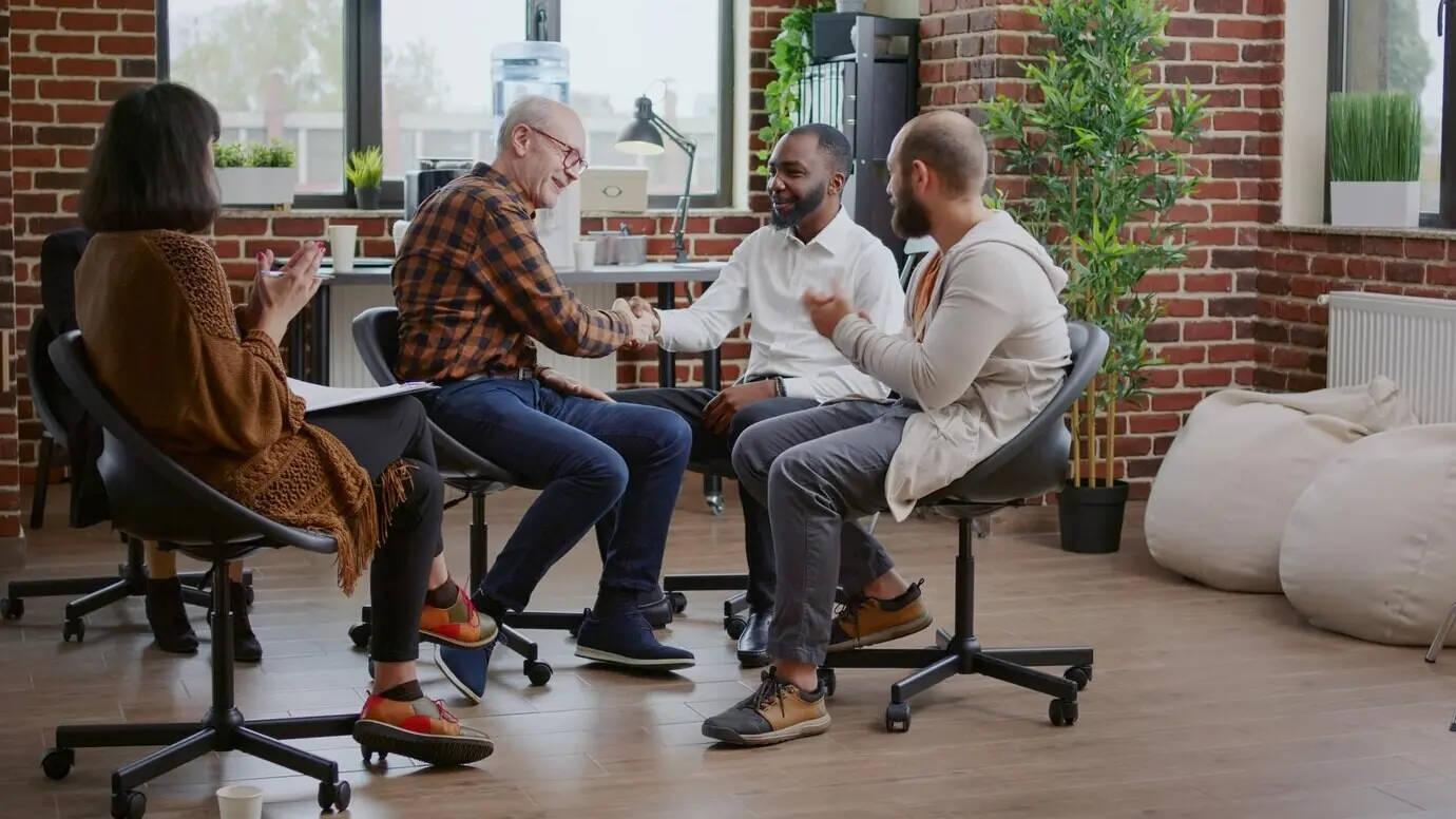 Hombre mayor recibiendo aplausos y estrechando la mano a personas en una reunión de un grupo de AA, celebrando el progreso. Persona compartiendo un logro terapéutico con el psicoterapeuta y los pacientes en rehabilitación.